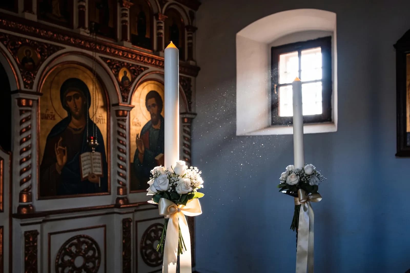 Două lumânări de cununie cu flori albe și panglici ivoire în fața iconostasului unei biserici ortodoxe, în lumina după-amiezii.